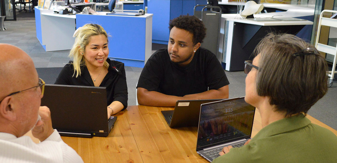 Three students and a teacher sitting around a table discussing the IT course