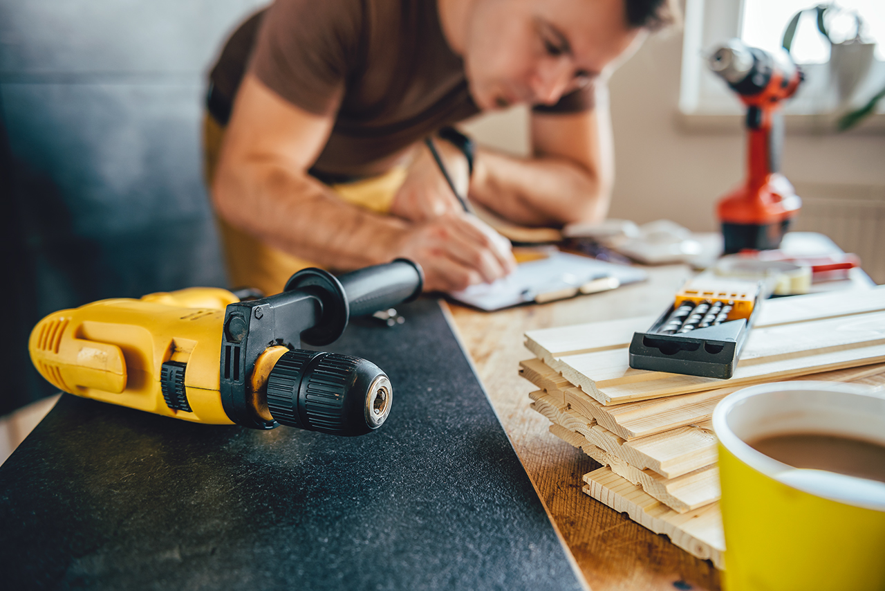 Yellow power drill and man making draft plan using pencil on the table with tools in the background